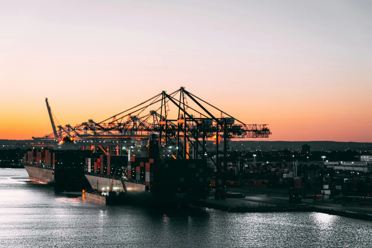Container ship at a working port at dusk, with cranes and operating lanes forming a compressed logistics system.
